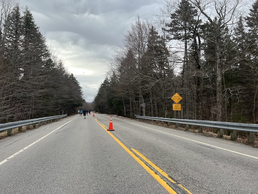Road with some runners in the distance and a warning sign that says "Moose Crossing" and "Next 8 Miles." 