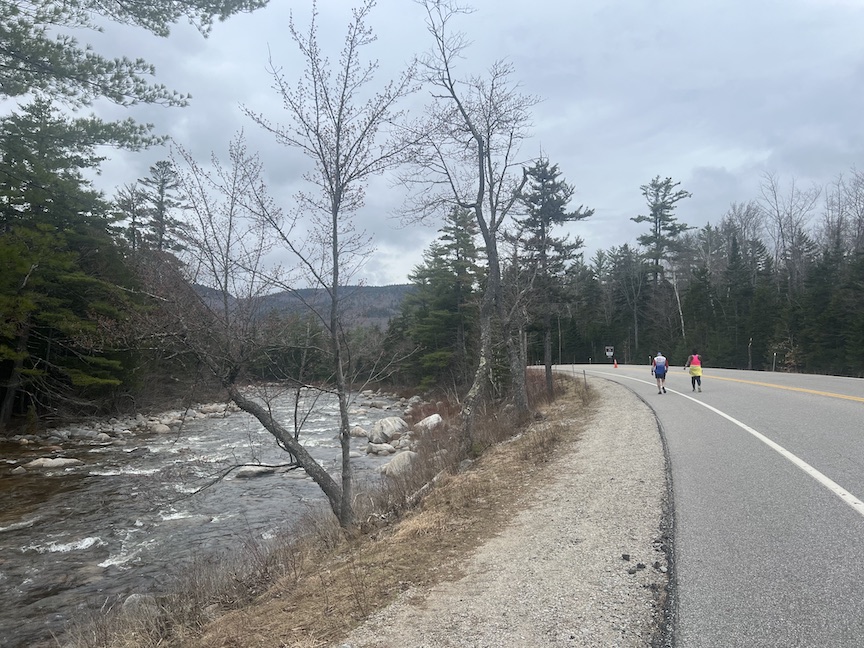 View from the REVEL White Mountains course on the Kancamagus Highway in New Hampshire. Two runners are in the distance. A rocky river is to the left and trees are around, with mountains in the distance. 