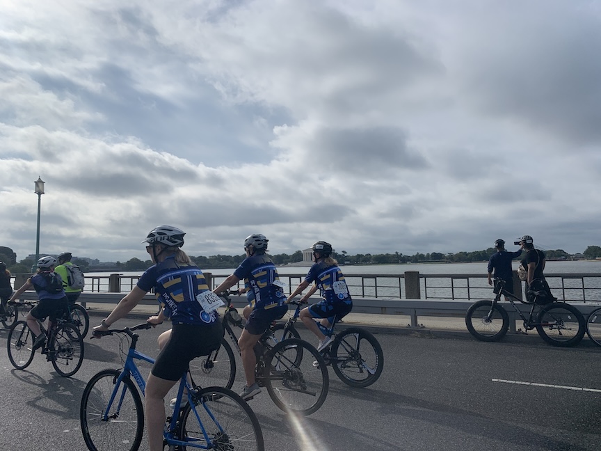 Cyclists ride past monuments during the DC Bike Ride. They are on a bridge, with water to their right. 