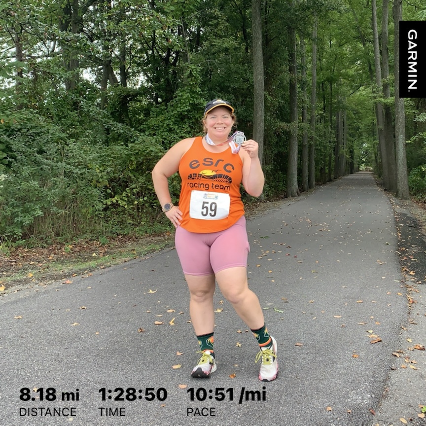 Vanessa Junkin poses with her medal on the Cross Island Trail, which has trees on both sides. Text at the bottom says 8.18 mi (distance), 1:28:50 (time), 10:51/mi (pace).