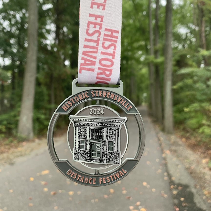 Close up of the finisher medal for the Historic Stevensville Distance Festival, with the Cross Island Trail in the background.