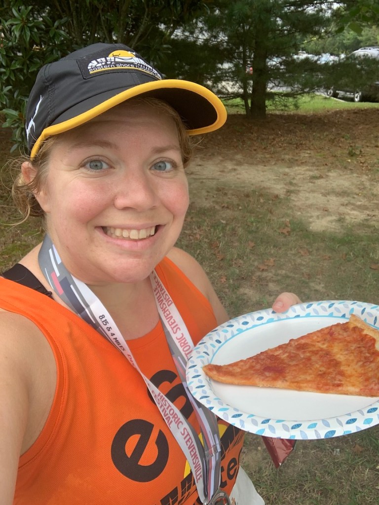 Vanessa Junkin, wearing running clothes and a medal, takes a selfie while holding a piece of pizza. 