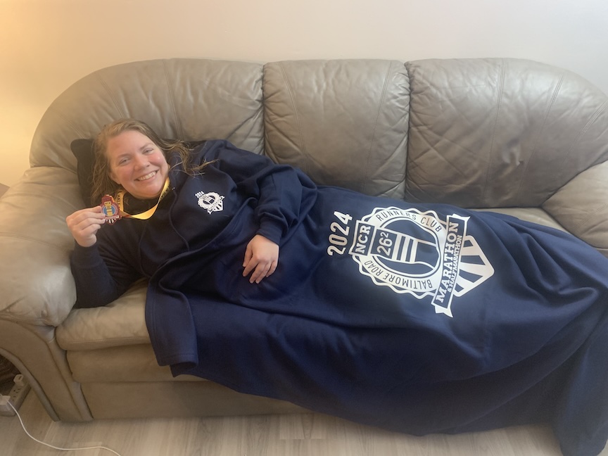 Runner relaxing on couch holding medal and wearing navy blue hoodie and under navy blanket with NCR Marathon logo on it. 