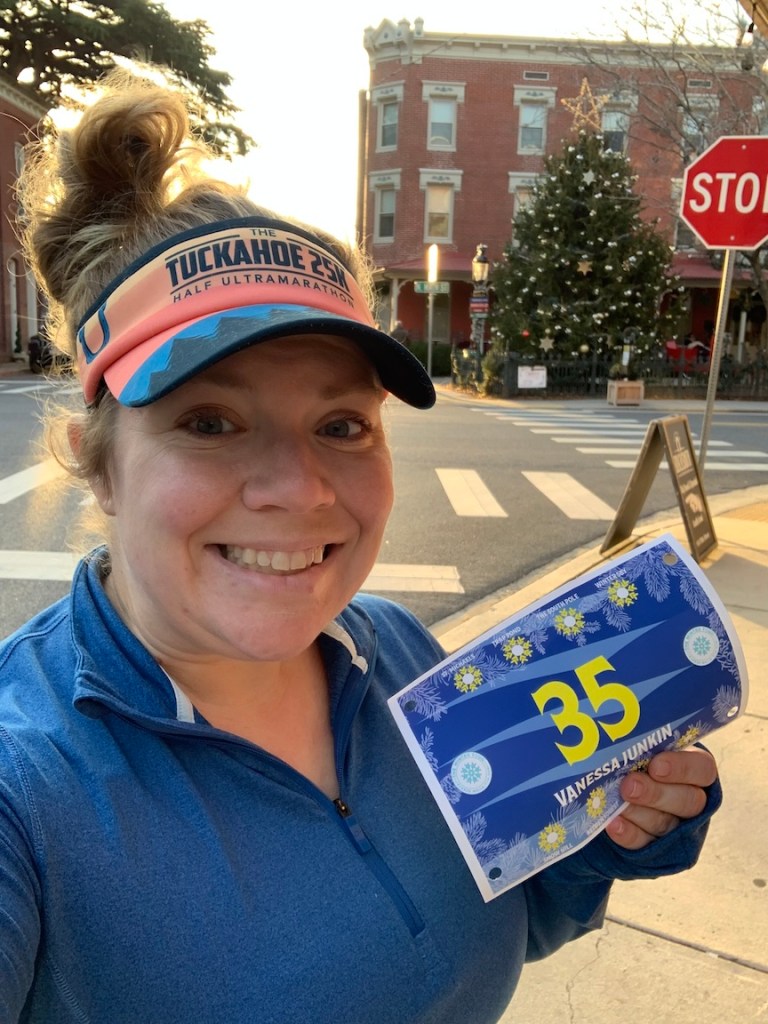 Selfie of Vanessa Junkin holding Winter Tour race bib with Christmas tree in the background. 