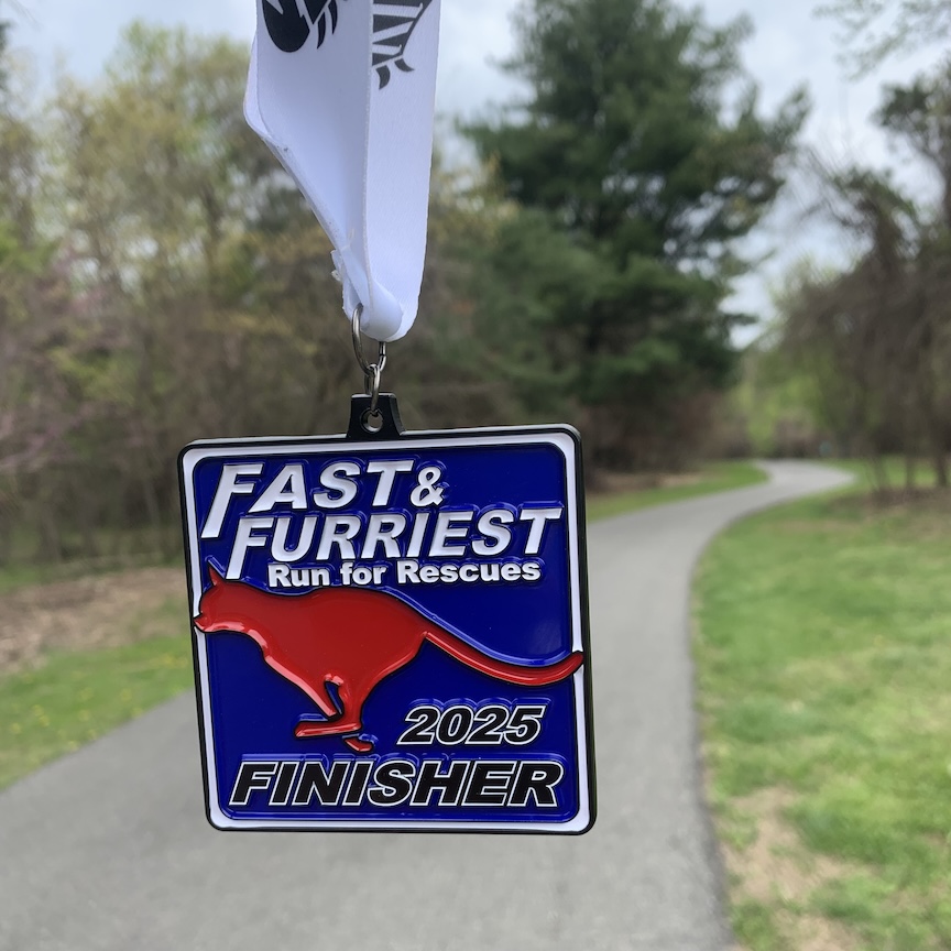 In the foreground is a Fast & Furriest Run for Rescues 2025 Finisher medal with a paved trail (the race course) in the background. 