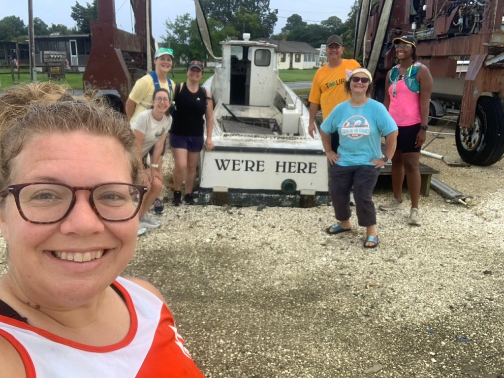 Group of runners posing by boat that says, "We're Here" on the back.