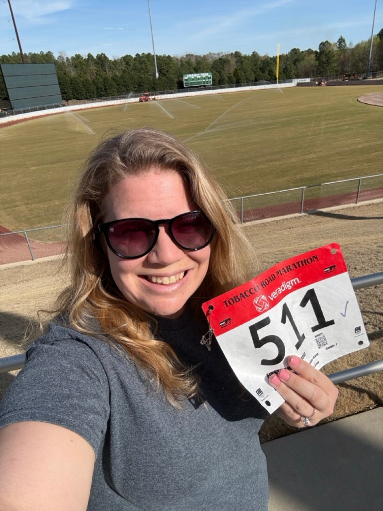 Selfie of Vanessa Junkin holding a race bib that says "511" with a baseball field in the background. 